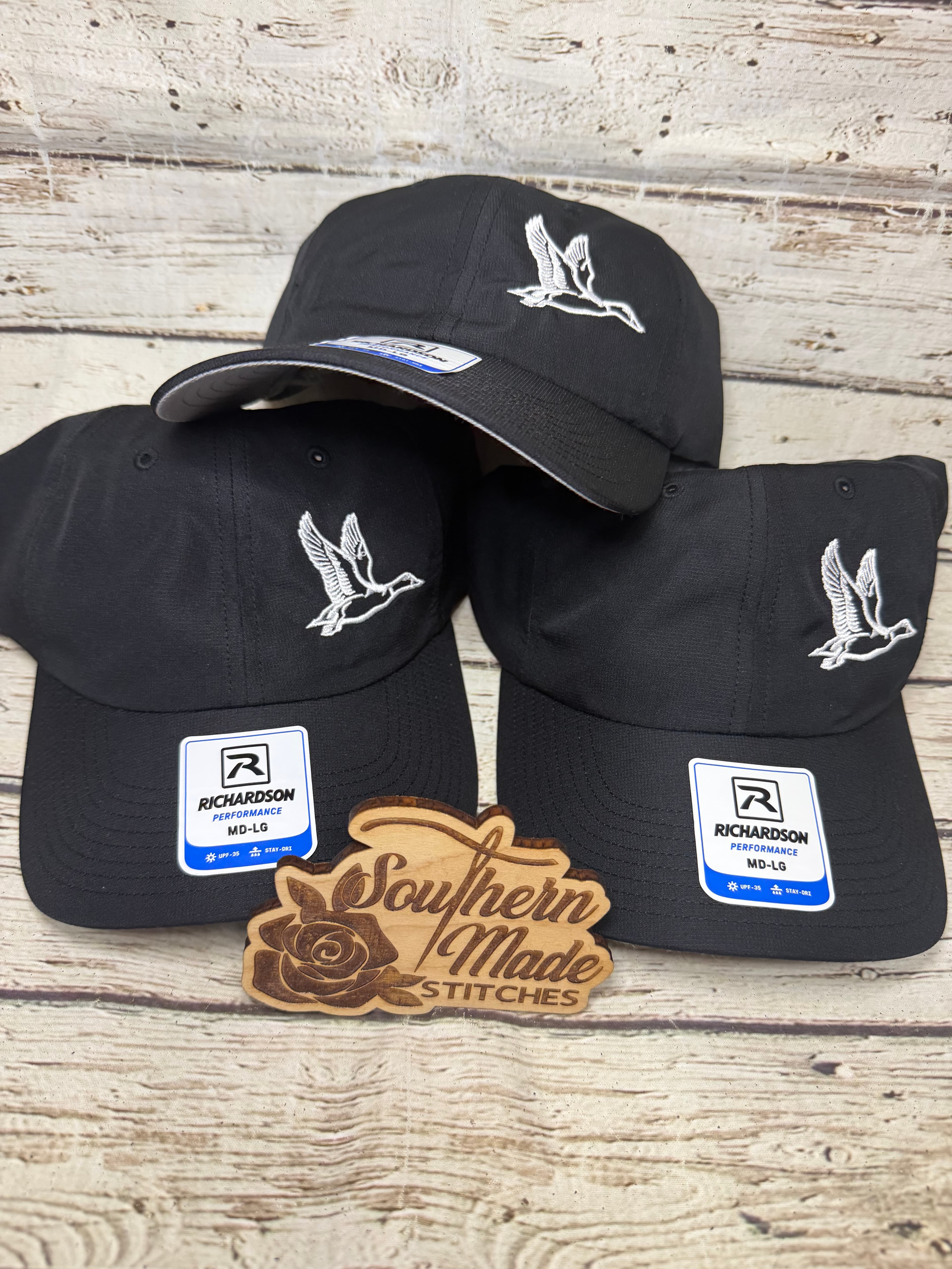 Three black Richardson performance caps featuring white embroidered flying duck logos on a wooden background.