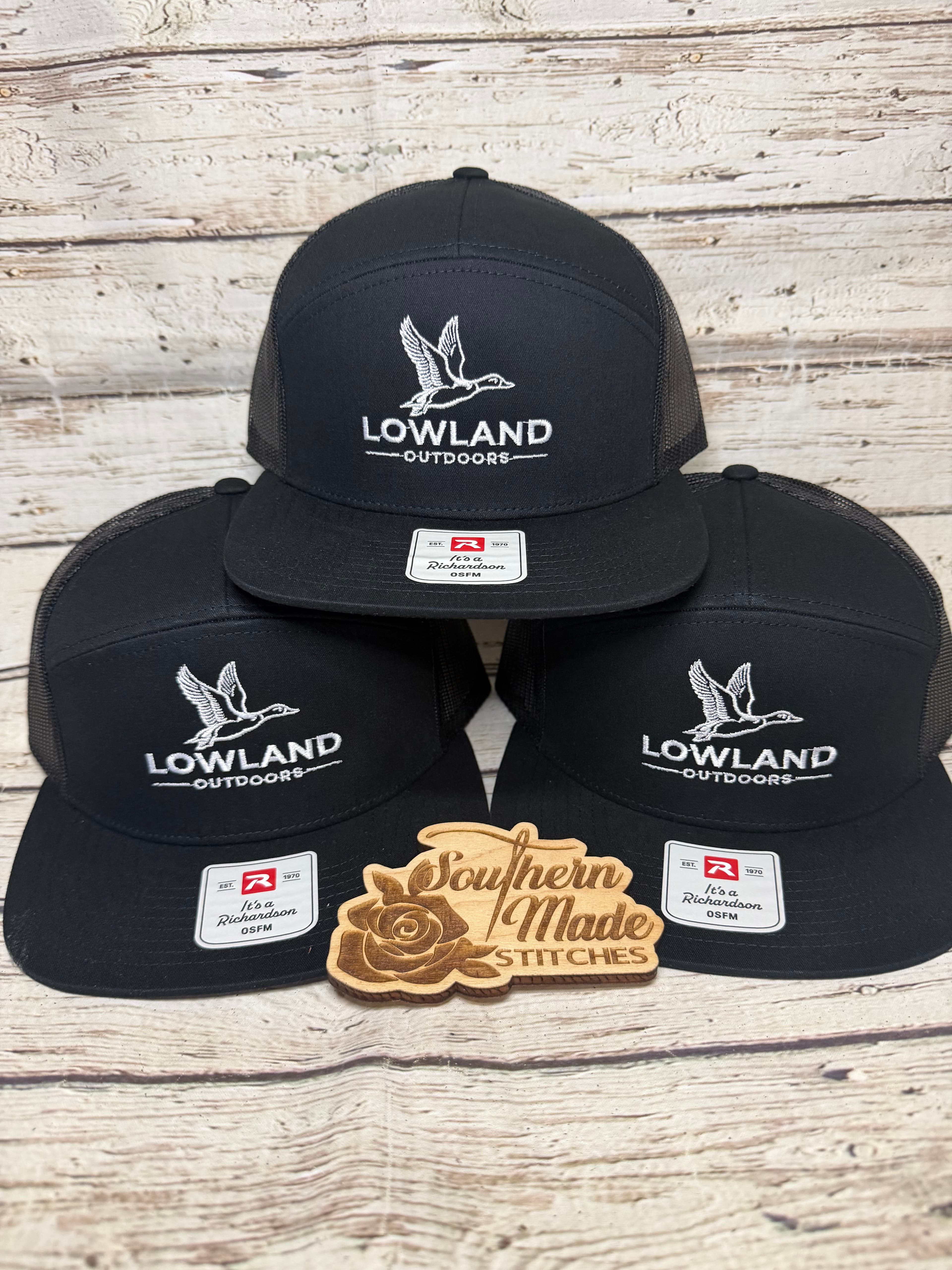 Three black hats with Lowland Outdoors duck logos and a Southern Made Stitches wooden sign.