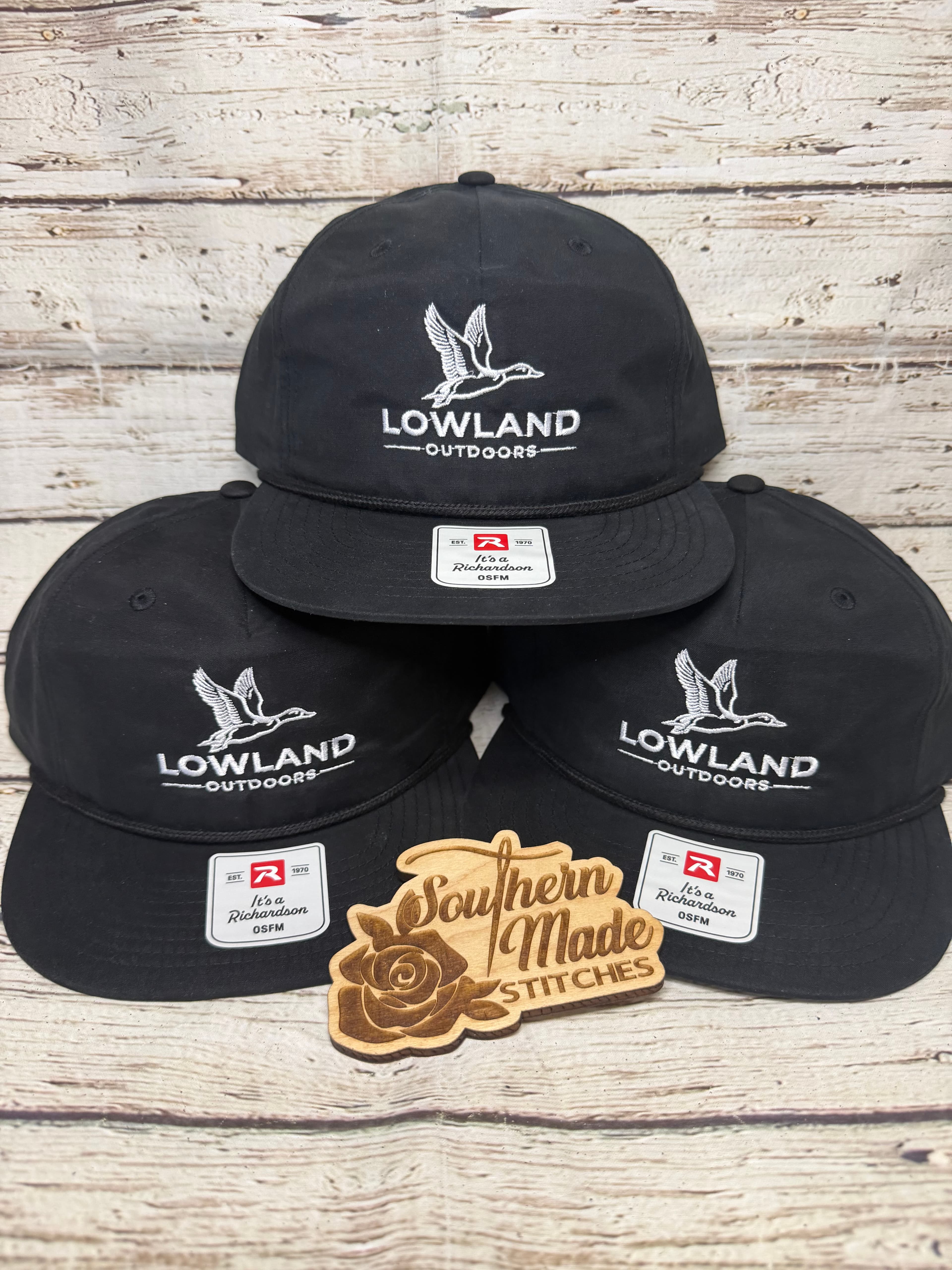 Three black hats with white Lowland Outdoors duck logos stacked on a wooden surface.